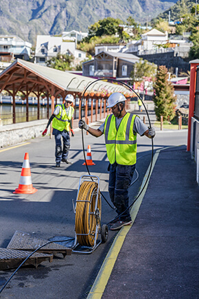 Deux techniciens en casque et gilet de sécurité déroulent un câble depuis une bobine sur une route, avec des cônes de signalisation autour de la zone de travail.