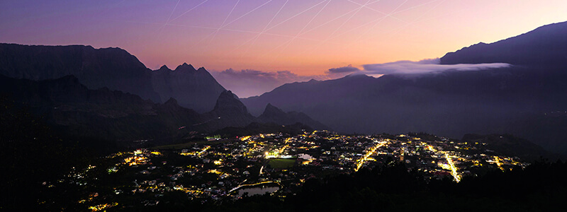 Village illuminé dans une vallée de montagne au crépuscule avec sommets escarpés, nuages bas et ciel rose orangé.
