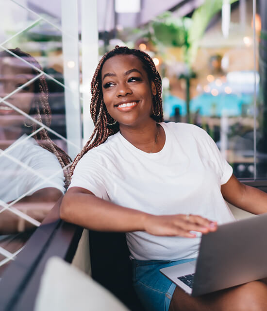 Une femme souriante assise près d’une fenêtre utilise un ordinateur portable dans un espace de travail moderne et lumineux.
