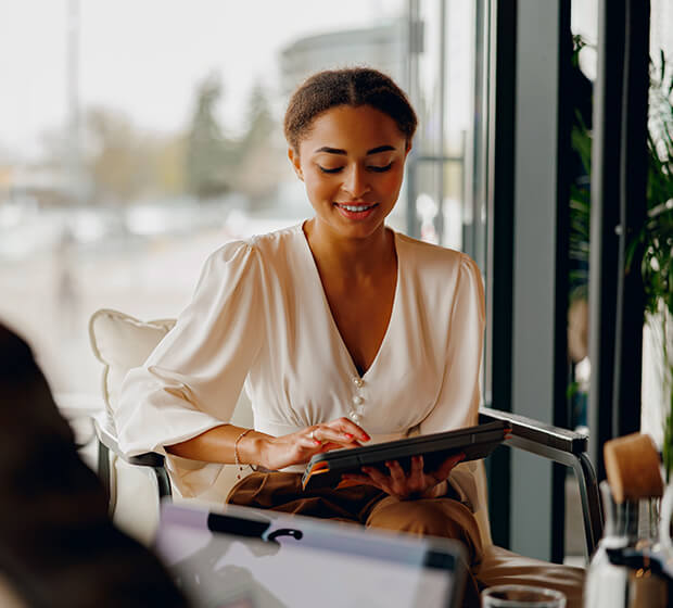 Une femme assise dans un intérieur lumineux consulte une tablette numérique, avec un environnement de bureau ou de salon en arrière-plan.