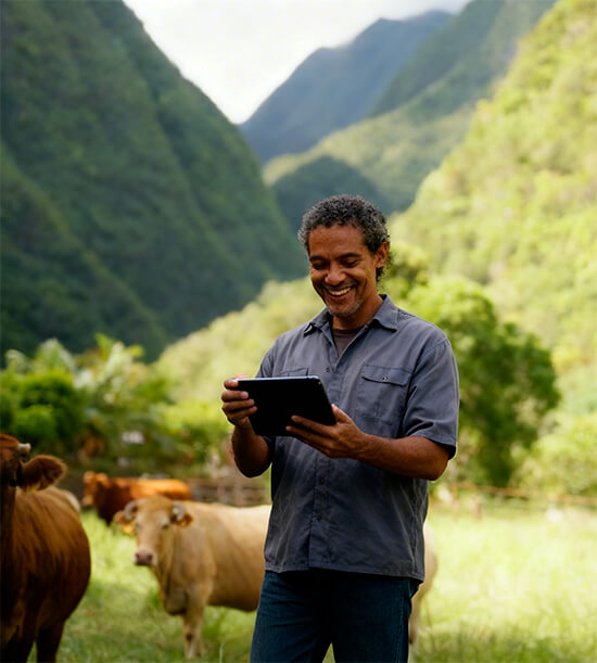 Un homme souriant utilise une tablette dans un pâturage avec des vaches, entouré de montagnes verdoyantes.